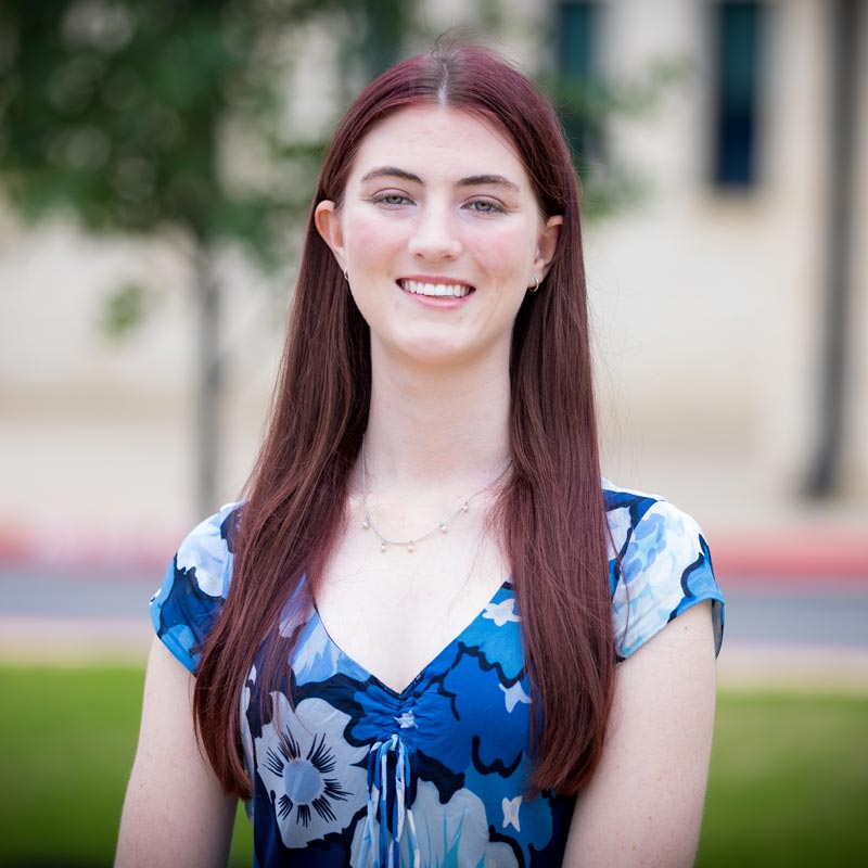 1CarolinePeterson26-sq Smiling woman with long red hair posing outside in a blue floral blouse.